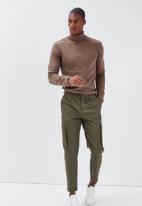 Brauner, enganliegender Rollkragenpullover kombiniert mit olivgrünen Cargo-Hosen mit Seitentaschen, ergänzt durch weiße Sneaker. Geschmeidige Textur, figurbetontes Design.