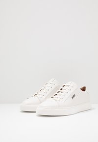 White leather sneakers with flat soles, round toe, and five eyelets. Features subtle branding on the side and a smooth texture.