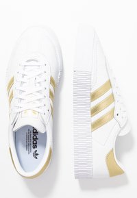 Un paio di sneakers bianche Adidas con strisce dorate, una scarpa vista dall'alto e l'altra di profilo, su uno sfondo bianco.