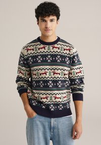 Suéter de punto en color crema con patrones de renos, árboles y copos de nieve multicolores. Puños y dobladillo de canalé en color navy. Combinado con jeans en un tono azul claro.