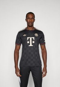Jersey negro de mangas cortas con un patrón de cuadros, detalles en blanco y un gran logo central. Presenta un ribete verde y rojo en el cuello y las mangas.