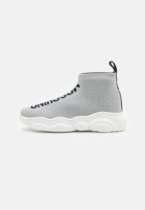 Šedá pletená kotníková bota s bílou gumovou podrážkou, zaoblenou špičkou a poutkem vzadu. Boční text v černé barvě.