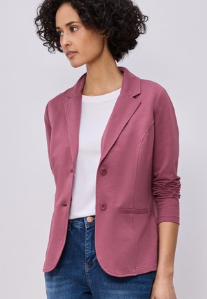 Vrouw met kort krullend haar, gekleed in een stofroze blazer met geplooide mouwen over een wit shirt en blauwe spijkerbroek, kijkt naar links.