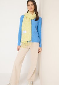 Écharpe à imprimé léopard vert clair avec des franges, portée sur un pull bleu. Un pantalon beige neutre et des baskets complètent la tenue.