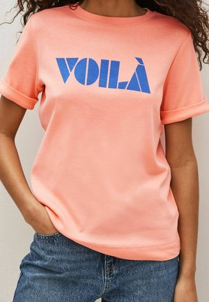 Femme portant un t-shirt corail clair avec des manches retroussées et un texte bleu "VOILÀ", associé à un jean bleu en denim, main dans la poche.