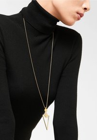 Gul geometrisk vedhæng halskæde med et trekantet design, hængende fra en tynd kæde, båret med en sort ribbet turtleneck top.