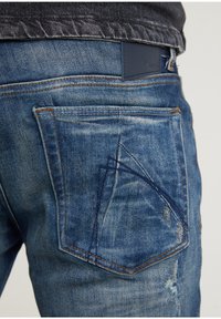 Farkut haalistuneessa sinisessä, joissa on teksturoitu viimeistely ja selkeä kolmion muotoinen kuvio takataskussa sekä oranssit ompeleet koristeena.