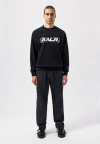 Sudadera negra con un logotipo blanco en la parte frontal, combinada con pantalones cargo oscuros. El modelo se encuentra frente a un fondo neutro, vistiendo zapatillas negras.