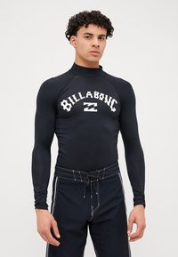 Czarna, długa koszulka rash guard z wysokim kołnierzem i białym logo "Billabong". Noszona z dopasowanymi czarnymi szortami pływackimi z białymi akcentami i sznurkiem.