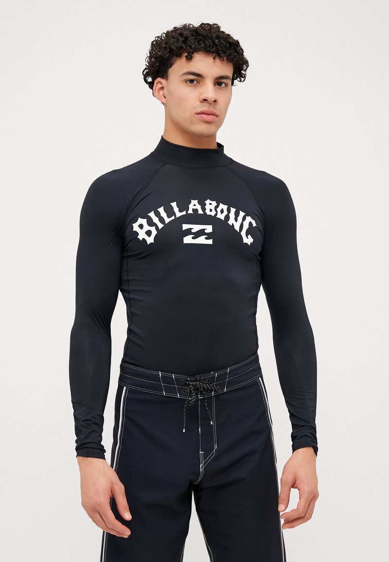 Melnas garas piedurknes rash guard krekls ar augstu apkakli un baltu "Billabong" logotipu. Valkāts ar saskanīgām melnām peldbiksēm, kurām ir baltas akcenti un aukla.