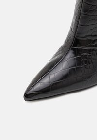 Bottine en cuir verni noir avec un bout pointu, présentant une texture en cuir de crocodile et une silhouette élégante. Aucun matériel ou accent visible.