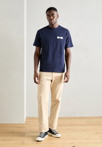 Námořnická modrá bavlněná tričko s bílým logem na prsou, spárované s béžovými kalhotami a černými teniskami s bílou podrážkou, stojící na dřevěné podlaze.