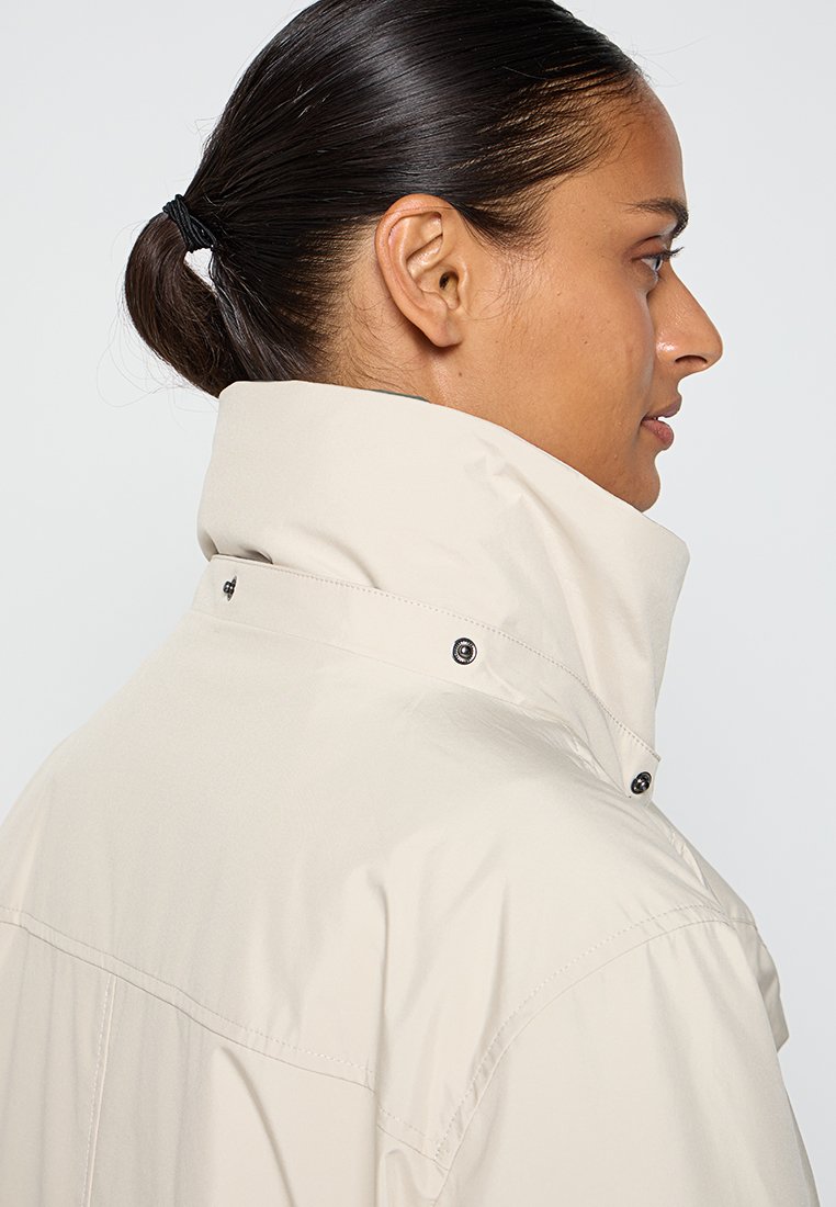 Profil d'une personne avec des cheveux bruns attachés, portant une veste beige clair à col haut avec des détails de boutons sur un fond blanc.