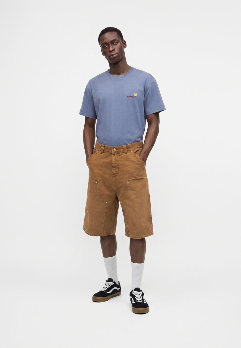 Hombre de pie con las manos en los bolsillos, lleva una camiseta azul, pantalones cortos marrones hasta la rodilla, calcetines blancos y zapatillas negras sobre un fondo liso.