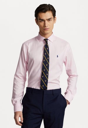 Lichtroze overhemddas met een button-down kraag, featuring een marineblauwe stropdas met diagonale strepen en embleem. Gecombineerd met marineblauwe broek met streepjes.