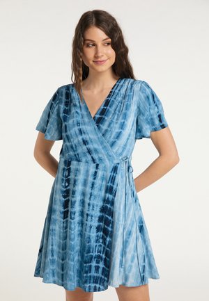Donna che indossa un vestito a maniche corte blu tie-dye con scollo a V, sorridente e che guarda di lato su uno sfondo chiaro e uniforme.