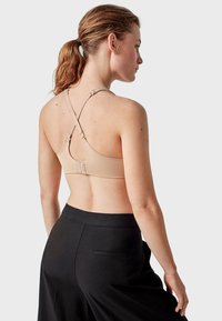 Soutien-gorge beige avec un design croisé dans le dos, des bretelles réglables et une fermeture à crochets. Porté avec un pantalon noir taille haute. Texture lisse.
