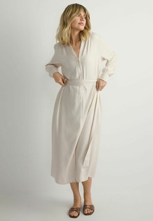 Kobieta poprawiająca wiązanie w talii beżowej sukienki midi z długim rękawem i guzikami, nosząca brązowe sandały z odkrytymi palcami na jednolitym tle.