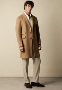 Hellbrauner Wollmantel mit tailliertem Schnitt, ausgestattet mit drei Knöpfen und zwei Seitentaschen, getragen über einem cremefarbenen Pullover und beigefarbenen Hosen, kombiniert mit braunen Schuhen.