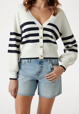 Femme portant un cardigan blanc boutonné avec des rayures noires et un short en jean bleu clair avec la main dans la poche.