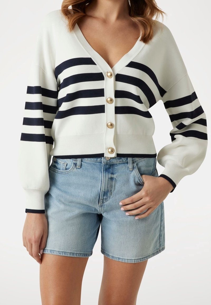 Femme portant un cardigan blanc boutonné avec des rayures noires et un short en jean bleu clair avec la main dans la poche.