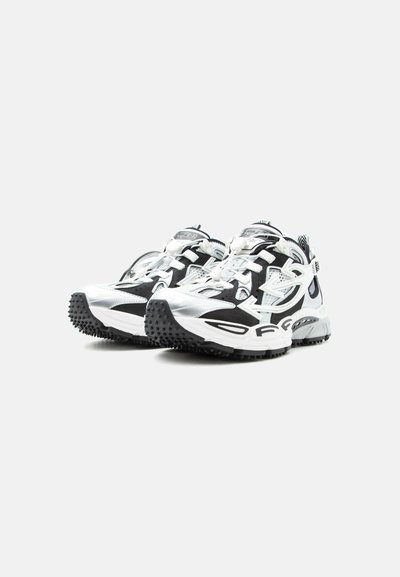 Χοντροειδή μαύρα και λευκά αθλητικά παπούτσια με ανθεκτικές, υφασμένες σόλες σχεδιασμένες για χρήση σε εξωτερικούς χώρους ή μονοπάτια, τοποθετημένα δίπλα το ένα στο άλλο σε φωτεινό φόντο.