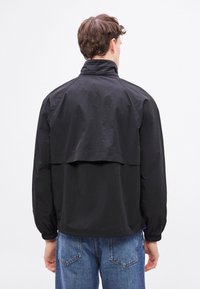 Jachetă neagră ușoară cu guler înalt, manșete elastice și design cu spate despărțit. Confecționată din material neted, rezistent la apă.