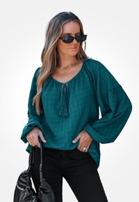 Teal textuurblouse met een brede halslijn en trekkoorddetail, gecombineerd met zwarte broek en een zwarte handtas. Lange, volumineuze mouwen.