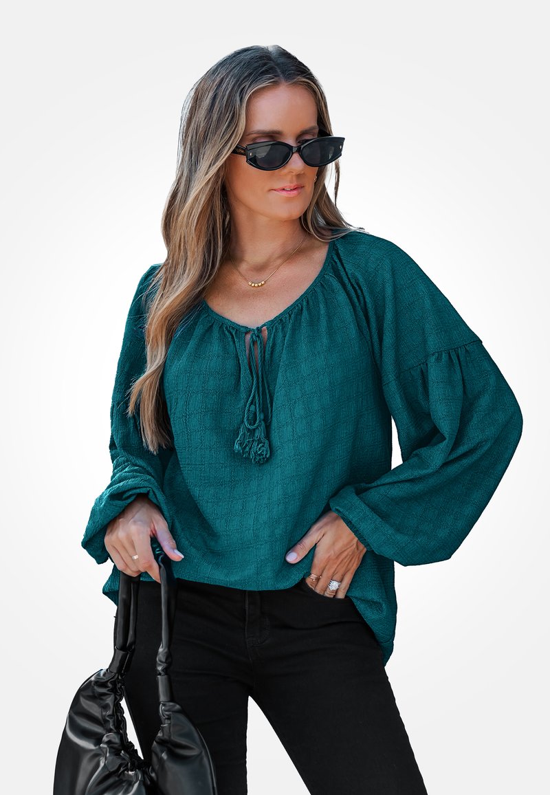 Teal textuurblouse met een brede halslijn en trekkoorddetail, gecombineerd met zwarte broek en een zwarte handtas. Lange, volumineuze mouwen.