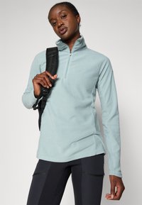 Pullover in pile azzurro chiaro con colletto a mezza zip, maniche lunghe e vestibilità rilassata. Indossato con pantaloni neri e una tracolla nera.