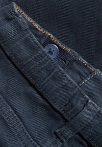 Tumma sininen farkkukangas, jossa näkyy ompelu ja vyötäröllä nappilisäys sekä litteä taskuyksityiskohta.