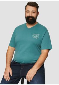 Teal v-hals t-shirt van katoen met korte mouwen. Bevat eenvoudige witte grafische tekst en een ontspannen pasvorm. Draagt met donkere jeans.