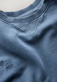 Niebieska bluza z teksturowanej tkaniny, z prążkowanym dekoltem i subtelnym wytłoczonym logo w lewym dolnym rogu.