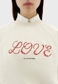 Cremefarbener Sweatshirt mit Raglanärmeln, verziert mit roter Schrift "LOVE" und schwarzem Text "ALL OR NOTHING" darunter. Glatte Textur.