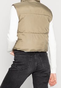 Světlě hnědá prošívaná vesta s vysokým límcem, nošená přes bílý žebrovaný svetr; doplněna tmavými džínami.