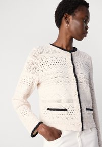 Cardigan lavorato a maglia in crema con motivi ad intaglio intricati, rifiniture nere, scollatura rotonda e chiusura a bottoni. Presenta due tasche con dettagli neri.
