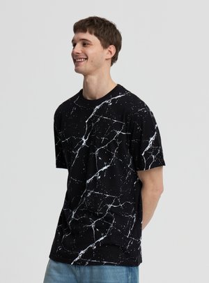 Giovane uomo sorridente che indossa una maglietta nera con motivo a marmo bianco e jeans azzurri chiari su sfondo grigio uniforme.