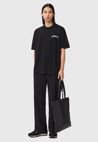 Sort oversize t-shirt med hvidt logo, sorte bukser med vide ben og en struktureret sort taske. Skoene er sorte med en lys sål.