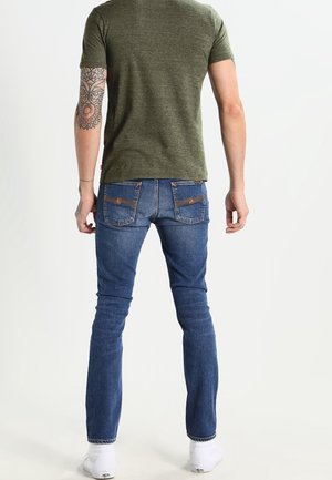 Hombre vistiendo una camiseta verde oliva, jeans azules con bolsillos traseros cosidos, calcetines blancos y un tatuaje visible en el antebrazo izquierdo, de pie de espaldas.