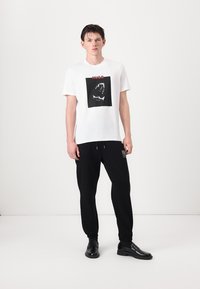 White cotton t-shirt with a black square graphic of a crocodile mouth, paired with black sweatpants featuring a small logo patch. Black shoes.
