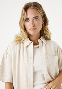 Kurzärmliges, beiges Hemd mit Knopfleiste aus strukturiertem Stoff, ausgestattet mit einer Brusttasche und einem lässigen Schnitt. Über einem weißen gerippten Oberteil getragen.