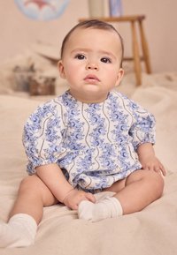 Fehér ruha kék virágmintával, pufós ujjakkal és gyűrt derékkal, fehér zoknival párosítva, semleges textúrájú felületen ülve.