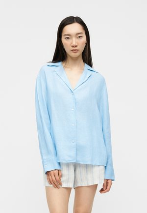 Young woman with long dark hair wearing a loose light blue button-up shirt and striped beige shorts, standing against a plain background.