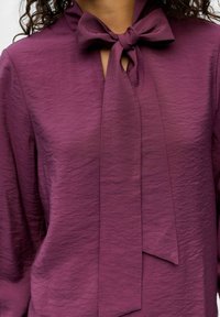 Lila Bluse aus gekräuseltem Stoff mit einer Schleife am Ausschnitt, langen Ärmeln und einer strukturierten Oberfläche.