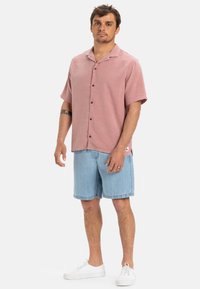 Różowa koszula na krótki rękaw z teksturowanej tkaniny, zestawiona z dżinsowymi szortami i białymi trampkami. Prosty kołnierzyk i dwa widoczne guziki.