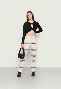 Camisola preta de mangas longas com um design em formato de buraco de chave, combinada com calças em tie-dye bege com riscas pretas. A modelo segura uma pequena bolsa preta.