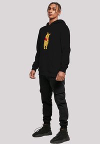 Zwarte hoodie met een gele cartoonbeer met een rood shirt op de voorkant, gemaakt van zacht materiaal en voorzien van een kangoeroezak.