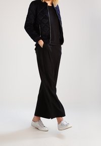 Marineblauer, gesteppter Bomberjacke, schwarzes Oberteil, schwarze weit geschnittene Hose und silberne Sneakers mit strukturierten Details. Das Model posiert mit den Händen in den Taschen.