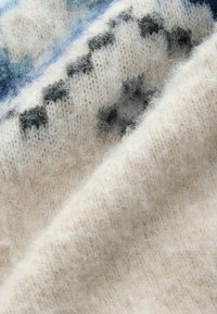 Tissu de pull en tricot en laine beige douce avec une texture duveteuse. Des accents bleus et noirs en motifs sont visibles dans le tissage.