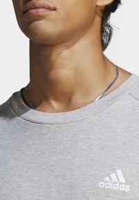 Siva trenirka z rebrastim ovratnikom, mehko teksturo in diskretnim logotipom Adidas v beli barvi na prsih. Ima rahlo sijočo površino.